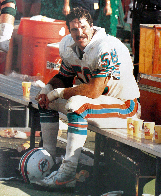 An old photo of Kim Bokamper sitting on the Miami Dolphins silver metal bench wearing his iconic 58 jersey, with his elbows resting on his knees looking left with a smile on his face. His wrists are wrapped with white tape and his hair is wet with sweat. Next to him on the bench are Wendy’s cups.