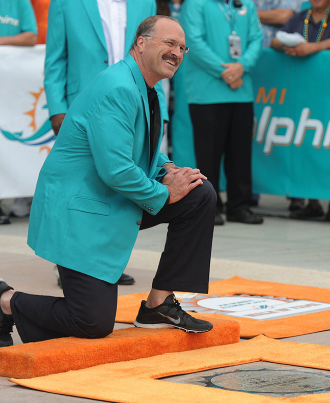 Photo shows kim ‘Bo’ Bokamper being inducted into the Miami Dolphins Walk of Fame. Wearing black pants, black Nike tennis shoes and a Miami Dolphins blue sports coat, Kim is kneeling on his right knee on an orange carpet in front of his induction plaque, hands on his left knee smiling. Background shows Miami Dolphins staff standing in front of the Miami Dolphins logos on fabric lining a fence with onlookers.